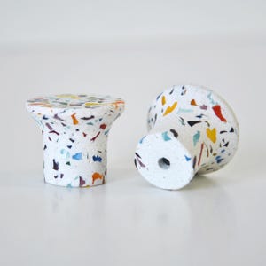Puede incluir: Dos pomos cilíndricos para armarios con base blanca y un patrón de terrazo multicolor con motas rojas, azules, amarillas y negras. Los pomos tienen una parte superior circular plana y un inserto de metal roscado visible en la base de uno.
