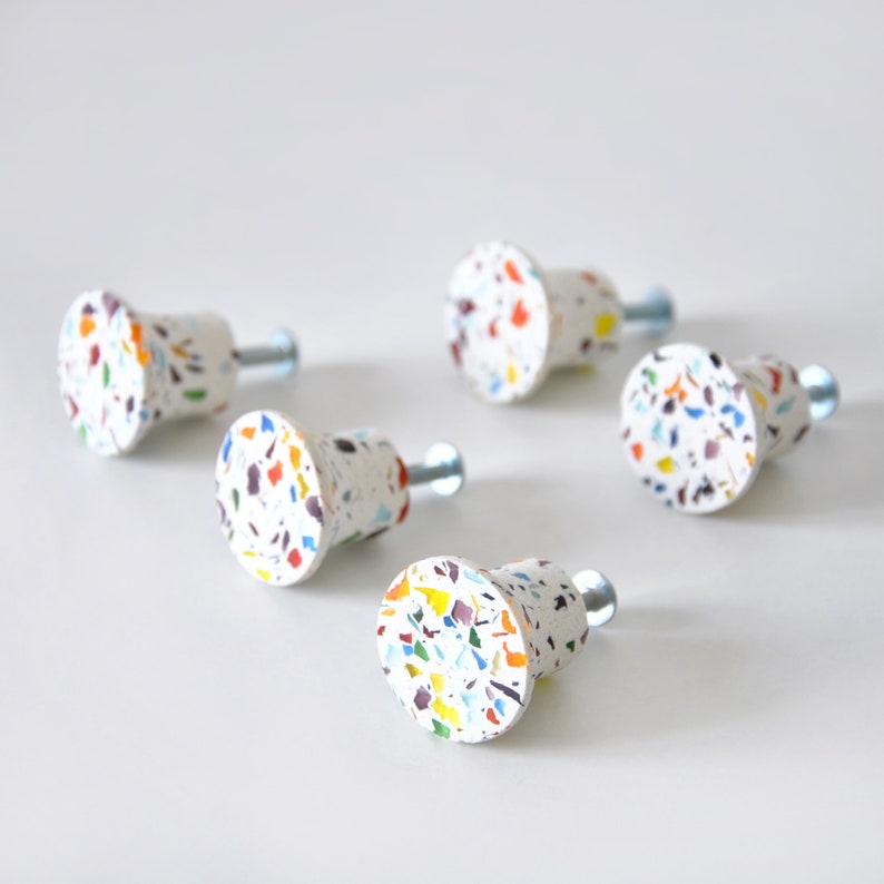 May include: A set of five cabinet knobs featuring a white base with a multi-colored terrazzo pattern of blue, yellow, orange, and dark red flecks. Each knob has a circular face and a silver-toned metal screw extending from the back for installation.