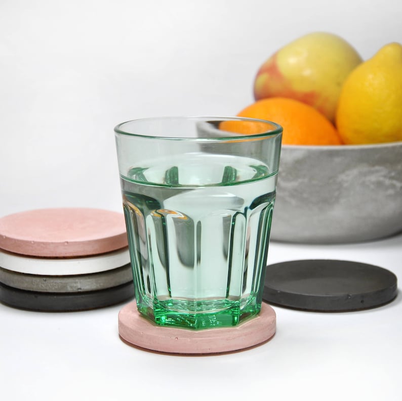 Puede incluir: Un vaso de agua sobre un posavasos de hormig&oacute;n rosa. El posavasos est&aacute; encima de una pila de otros cuatro posavasos en negro, gris, blanco y rosa. Hay fruta en un cuenco en el fondo.