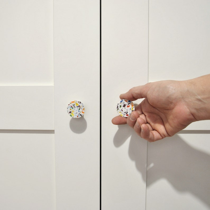 May include: Two circular cabinet knobs with a white base and a multi-colored terrazzo pattern featuring flecks of blue, yellow, orange, and black. The knobs are mounted on a plain white cabinet door with a vertical seam running down the center.