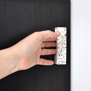 Può includere: Una maniglia per armadio in terrazzo bianco, nero e marrone. La maniglia è rettangolare e ha una finitura liscia e lucida.