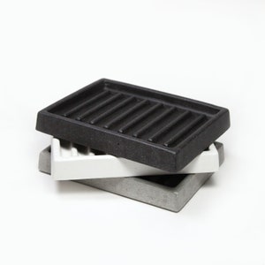 Può includere: Tre portasapone impilati uno sopra l'altro. Il portasapone superiore è nero con una superficie rigata. Il portasapone intermedio è bianco e il portasapone inferiore è grigio.