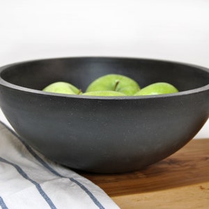 May include: A black ceramic bowl filled with green apples. The bowl is sitting on a wooden surface with a white and blue striped cloth.