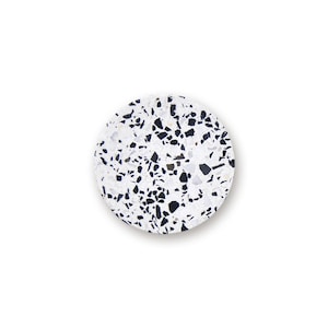 May include: A circular coaster featuring a white base with a scattered pattern of black and dark gray angular stone chips. The surface has a speckled terrazzo appearance with a smooth finish and a clean, defined edge against a plain white background.