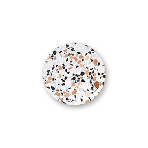 May include: A circular coaster featuring a white base with a scattered terrazzo pattern. The surface is embedded with various angular chips in shades of black, light gray, and warm terracotta brown, creating a multi-colored speckled appearance.