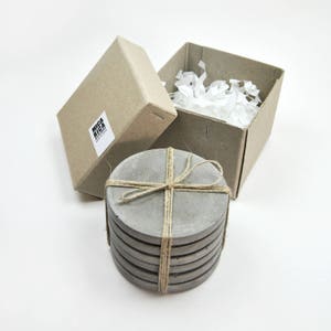 Puede incluir: Conjunto de seis posavasos de hormig&oacute;n gris atados con cuerda. Los posavasos est&aacute;n empaquetados en una caja de cart&oacute;n marr&oacute;n con las palabras "Mica Rica" impresas en el lateral.