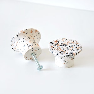 Könnte beinhalten: Zwei weiße Keramik-Schrankknöpfe mit einem Terrazzo-Muster in Schwarz, Braun und Weiß. Die Knöpfe haben eine runde Oberseite und eine leicht ausgestellte Basis.