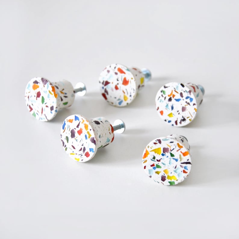 May include: Five round cabinet knobs featuring a white base with a multi-colored terrazzo pattern of small red, blue, yellow, and black flecks. Each knob has a short metal screw extending from the back for attachment to furniture or cabinetry.