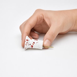 May include: A white cabinet knob with a speckled terrazzo pattern featuring small red and grey flecks. The knob has a flared, cylindrical shape and includes a gold-colored metal screw protruding from the back for installation purposes.
