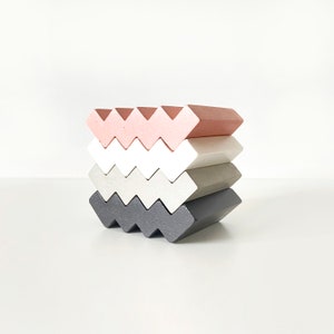 Può includere: Un set di cinque sottobicchieri di forma geometrica in rosa, bianco, grigio e nero. I sottobicchieri sono realizzati in un materiale liscio e testurizzato e hanno un motivo a zig-zag.