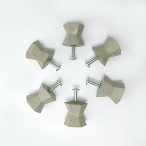May include: Six gray concrete cabinet knobs with silver screws arranged in a circular pattern. The knobs have a geometric, hourglass-like shape, and are designed for furniture or decorative use.