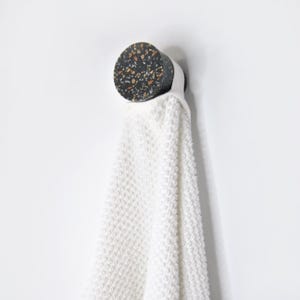 May include: A white towel hangs from a round black and brown terrazzo hook mounted on a white wall.