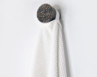 May include: A circular wall hook featuring a dark gray terrazzo pattern with flecks of orange, white, and black. A white, textured, waffle-weave towel hangs from the hook, draping downward in a soft, folded vertical orientation.