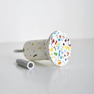 May include: A white cabinet knob with colourful terrazzo-style accents. The knob has a cylindrical body and a round face. A silver screw and a grey wall anchor are next to the knob.