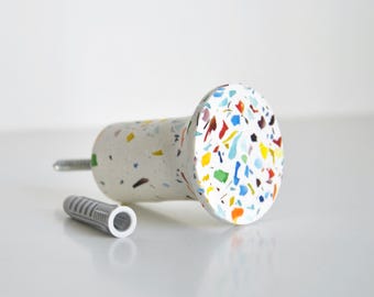 May include: A white cabinet knob with colorful terrazzo-style accents. The knob has a cylindrical body and a round face. A silver screw and a gray wall anchor are next to the knob.