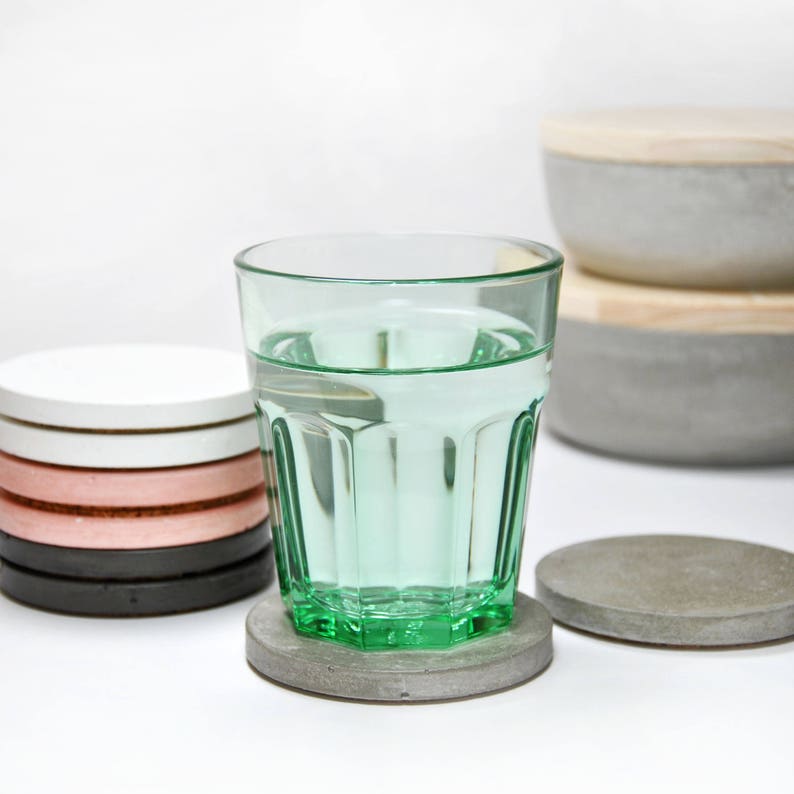 Puede incluir: Un vaso de vidrio verde con agua se encuentra sobre un posavasos de hormig&oacute;n gris. El posavasos est&aacute; sobre una superficie blanca. Hay cuatro posavasos redondos apilados en el fondo, con colores blanco, rosa, negro y gris.
