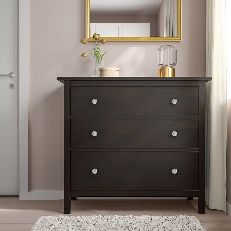 May include: A dark brown wooden dresser featuring three horizontal drawers, each equipped with two round, light-colored decorative knobs. The dresser has a rectangular frame with four sturdy legs and a flat top surface for storage or display.