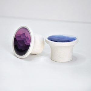 Op de afbeelding: Twee decoratieve kastknoppen. Eén knop heeft een paars bergketenontwerp, de andere een blauw-wit ontwerp. Beide knoppen hebben een witte, getextureerde basis en een afgeronde bovenkant.