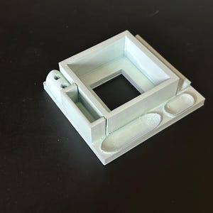 May include: A light green 3D printed model of a small building with a square window and two small rectangular openings on the side. The model is sitting on a black surface.