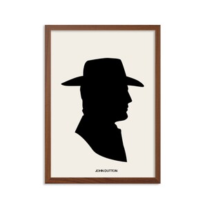 Op de afbeelding: Ingelijste kunstdruk met een zwart silhouet van een persoon met hoed op een crèmekleurige achtergrond, in een bruine houten lijst. De tekst "JOHN DUTTON" is in het zwart onderaan gedrukt. Minimalistisch ontwerp voor interieurdecoratie.