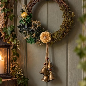 May include: A circular wreath made of brown woven twigs, decorated with green moss, dried foliage, and pale yellow flowers. Four brass-coloured metal bells hang from the bottom of the wreath, suspended by thin jute twine with small brown wooden beads.