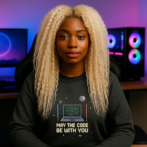 Könnte beinhalten: Eine Person trägt ein dunkelgraues Sweatshirt mit einer Grafik eines Laptops mit Binärcode, einem Mond und zwei strahlartigen Balken. Der Text "MAY THE CODE BE WITH YOU" steht darunter. Die Person hat lange, lockige blonde Haare.