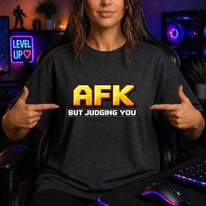 Könnte beinhalten: Ein dunkelgraues T-Shirt mit verpixeltem gelbem Text, der AFK lautet, und darunter weißem verpixeltem Text, der BUT JUDGING YOU lautet. Das Design ist mittig auf der Brust des T-Shirts platziert, das einen Rundhalsausschnitt und kurze Ärmel hat.