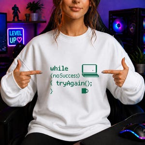 Könnte beinhalten: Weißes Sweatshirt mit grünem pixeligem Code, einem Laptop-Symbol und dem Text "while (noSuccess) { tryAgain(); }". Ein lässiges Kleidungsstück, geeignet für den Alltag.