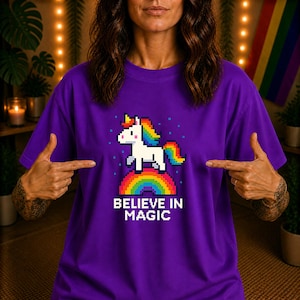 Könnte beinhalten: Ein lila T-Shirt mit einem pixeligen Einhorn-Design. Das Einhorn hat eine Regenbogenmähne und -schwanz und steht auf einem Regenbogen. Der Text "BELIEVE IN MAGIC" steht darunter. Das T-Shirt hat eine einfarbige lila Farbe.