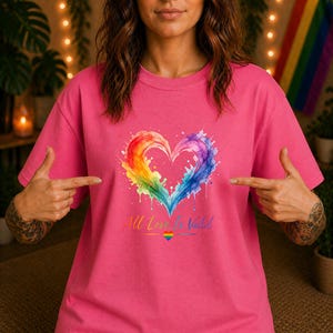 Könnte beinhalten: Ein leuchtend pinkfarbenes T-Shirt mit einem großen, aquarellartigen Herzmotiv in Regenbogenfarben. Unter dem Herz steht der Text All Love Is Valid in Schreibschrift, ergänzt durch ein kleines, mittig platziertes Regenbogen-Herz-Symbol.