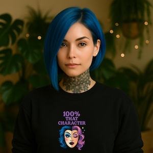 Könnte beinhalten: Eine Person mit blauen Haaren und einem schwarzen Sweatshirt. Auf dem Sweatshirt steht der Text "100% THAT CHARACTER" über einer geteilten Gesichts-Grafik mit blauen und rosa Haaren. Die Person hat ein Hals-Tattoo und schaut in die Kamera.