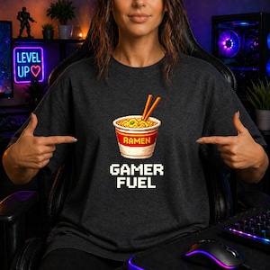 Könnte beinhalten: Ein dunkelgraues T-Shirt mit einer Pixel-Art-Grafik eines Bechers Ramen-Nudeln mit Essstäbchen. Unter dem Becher steht der Text GAMER FUEL in einer weißen, verpixelten Schriftart, der mittig auf der Brust des Kleidungsstücks platziert ist.
