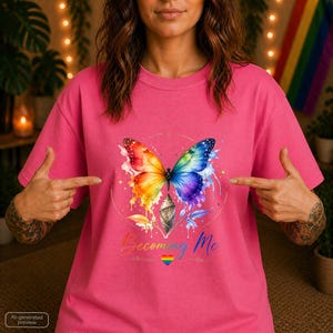 Könnte beinhalten: Ein leuchtend pinkfarbenes T-Shirt mit einer Aquarell-Grafik eines Schmetterlings, dessen regenbogenfarbene Flügel aus einer Puppe hervorgehen. Unter dem Schmetterling steht der Text Becoming Me in Schreibschrift, ergänzt durch ein kleines Regenbogenherz.