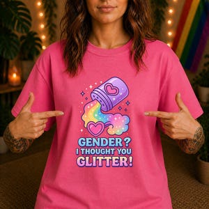 Könnte beinhalten: Ein pinkfarbenes T-Shirt mit einem farbenfrohen Grafikdesign. Das Design zeigt einen lila Behälter, der einen Regenbogen, Herzen und den Text "GENDER? I THOUGHT YOU GLITTER!" ausgießt.