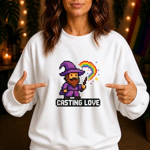 Könnte beinhalten: Weißes Sweatshirt mit einem pixeligen Zauberer in lila Roben und Hut, der einen Zauberstab mit einem Regenbogenbogen hält. Der Text "CASTING LOVE" steht darunter.