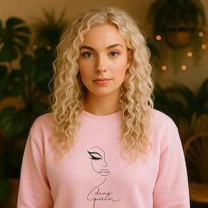 Könnte beinhalten: Hellrosa Sweatshirt mit schwarzer Strichzeichnung eines Gesichts und dem Schriftzug "drag queen" in Schreibschrift. Das Sweatshirt hat einen Rundhalsausschnitt und ist aus weichem Material.