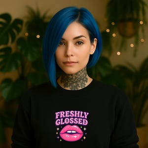 Könnte beinhalten: Ein schwarzes Sweatshirt mit dem Text "FRESHLY GLOSSED" in Rosa und Weiß, über einer rosa Lippen-Grafik. Die Person auf dem Bild hat blaue Haare und ein Tattoo am Hals.