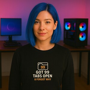 Könnte beinhalten: Eine Person trägt ein schwarzes Sweatshirt mit dem Text "GOT 99 TABS OPEN & FORGOT WHY" und einer Ordnergrafik mit der Nummer 99. Ein Computermonitor und ein Desktop-Computer sind im Hintergrund zu sehen.