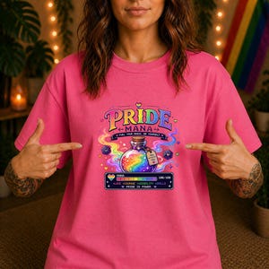 Könnte beinhalten: Ein leuchtend pinkes T-Shirt mit einem Grafikdruck einer Zaubertrankflasche mit regenbogenfarbener Flüssigkeit, umgeben von bunten Wirbeln und Würfeln. Der Text lautet PRIDE MANA, FUEL YOUR MAGIC, BE YOURSELF und PRIDE IS POWER, mit einer Manaleiste.
