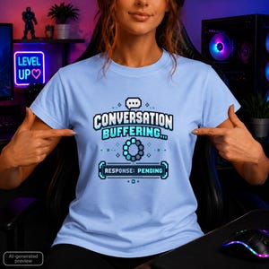 Könnte beinhalten: Ein hellblaues T-Shirt mit einem verpixelten Grafikdesign, das den Text "CONVERSATION BUFFERING..." über einem Ladekreis-Symbol und einem rechteckigen Kasten mit dem Text "RESPONSE: PENDING" in einem digitalen, retro-inspirierten Schriftstil zeigt.