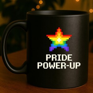 Könnte beinhalten: Ein mattschwarzer Keramikbecher mit einer verpixelten, regenbogenfarbenen Sterngrafik und einem kleinen weißen Pluszeichen in der Mitte. Unter dem Stern ist der Text PRIDE POWER-UP in einer weißen, blockartigen Pixel-Schriftart auf der Vorderseite gedruckt.