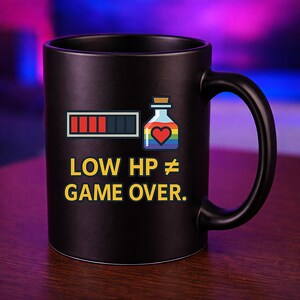 Könnte beinhalten: Ein mattschwarzer Keramikbecher mit einer Grafik, die eine Gesundheitsleiste mit roten Segmenten, eine regenbogenfarbene Zaubertrankflasche mit einem roten Herzsymbol und den gelben Text LOW HP ≠ GAME OVER zeigt. Der Becher hat einen Henkel.