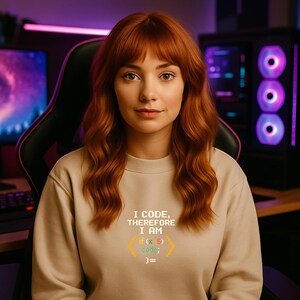 Peut inclure: Un sweat-shirt beige avec le texte "I CODE, THEREFORE I AM" et des extraits de code en jaune et vert. La personne sur l'image a les cheveux roux et est assise dans une chaise de jeu.