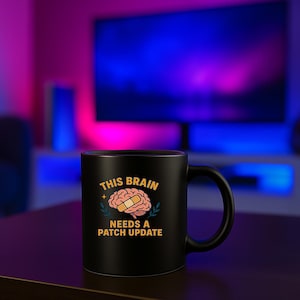Könnte beinhalten: Schwarze Keramik-Tasse mit dem Text "THIS BRAIN NEEDS A PATCH UPDATE" und einer Gehirn-Grafik mit Pflaster. Die Tasse hat einen gebogenen Henkel und steht auf einer dunklen Oberfläche.