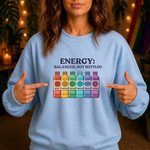 Könnte beinhalten: Hellblaues Sweatshirt mit dem Text "ENERGY: BALANCED, NOT BOTTLED". Darunter befinden sich sechs farbige Flaschen mit den Wörtern "ROOT", "SACRAL", "SOLAR", "THROAT" und "CROWN". Jede Flasche hat ein Energiesymbol.