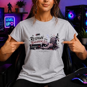 Könnte beinhalten: Ein hellgraues T-Shirt mit einem Grafikdruck einer Kapuzenfigur, die neben einem Grabstein mit der Aufschrift Game Over liegt. Der Text lautet Respawn Tomorrow, You've done your best. Come back tomorrow und Try again tomorrow mit Batteriesymbol.