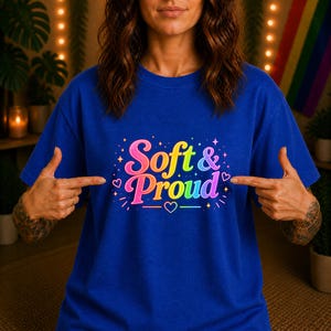 Könnte beinhalten: Ein königsblaues T-Shirt mit dem Schriftzug Soft & Proud in einer mehrfarbigen Regenbogen-Schriftart. Das Design ist mit kleinen Herzen und Sternen verziert, die den Text umgeben und eine lebendige, farbenfrohe Grafik bilden.