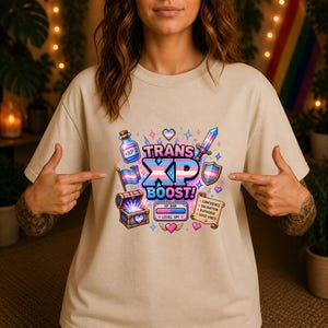 Könnte beinhalten: Ein beiges T-Shirt mit einer bunten Pixel-Art-Grafik und dem Text "TRANS XP BOOST!". Das Design zeigt einen Trankflakon, ein Schwert, einen Schild, eine Schatztruhe, eine Schriftrolle mit "CONFIDENCE, VALIDATION, EUPHORIA, GOOD VIBES" und Flaggen.
