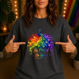 Könnte beinhalten: Ein anthrazitfarbenes T-Shirt mit einem lebendigen Pfau im Aquarellstil, dessen Schwanzfedern in Regenbogenfarben leuchten. Unter dem Pfau steht der Text "Be Proud. Be You." sowie der Zusatz "BEAUTIFUL IN EVERY COLOR" in schlichter Schrift.