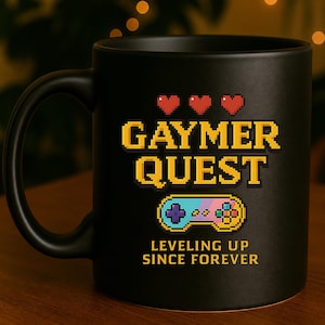 Könnte beinhalten: Schwarze Keramik-Tasse mit dem Text "GAYMER QUEST" in gelber Pixel-Schrift, einem pixeligen Gamecontroller, drei roten Herzen und dem Satz "LEVELING UP SINCE FOREVER".